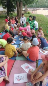 Grupa dzieci siedząca na matach, na trawie słuchająca czytanych wierszy i kolorująca kolorowanki. Na drugim planie uczniowie liceum czytający wiersze.