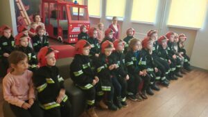 Grupa dzieci w kurtkach i hełmach strażackich.