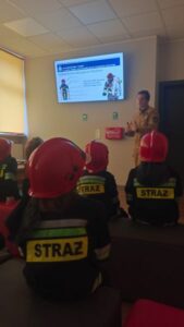 Grupa dzieci w hełmach i kurtkach strażackich ogląda prezentację.