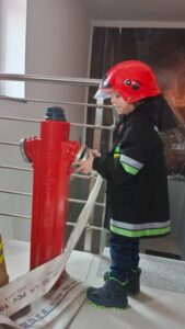Chłopiec w czerwonym hełmie i kurtce strażackiej stoi przy hydrancie.
