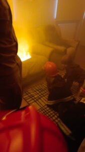 Dzieci w hełmach strażackich w zadymionym pomieszczeniu. W tle ogień wydobywający się z kanapy.
