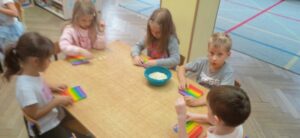 Dzieci siedzą przy stoliku na którym stoi miseczka z fasolkami, każdez nich ma przed sobą pop-it.