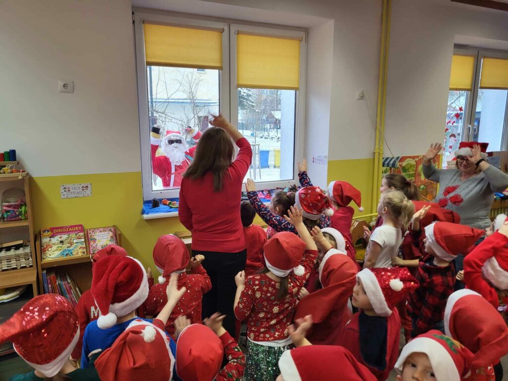 dzieci i nauczycielki w czerwonych strojach machające św. Mikołajowi zaglądającemu przez okno