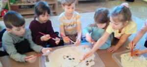 Grupa dzieci przy pomocy pędzelków odkrywa kamienie i odciski z masy solnej znajdujące się na tackach z kaszą.