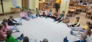 Grupa dzieci siedzi w kole na dywanie, wszyscy trzymają gumę sensoryczną.