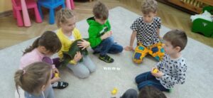 Grupa dzieci siedzi na dywanie, przed nimi znajdują się kartoniki z literami i plastikowe żółte jajko.