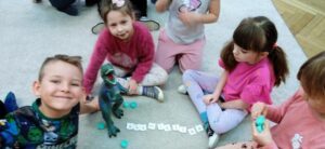 Grupa dzieci siedzi na dywanie, przed nimi znajdują się kartoniki z literami i plastikowe zielone jajka, figurka dinozaura.