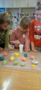 Dzieci stoją przy stoliku na kórym znajduje się tacka z kolorowymi jajkami uformowanymi z masy. Jedno dziecko wkłada pipetę do białego kubeczka.