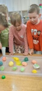 Dzieci stoją przy stoliku, na którym znajduje się tacka z kolorowymi jajkami uformowanymi z masy sodowej.