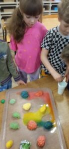 Dzieci stoją przy stoliku na kórym znajduje się tacka z kolorowymi jajkami uformowanymi z masy. Jedno dziecko wkłada pipetę do białego kubeczka.