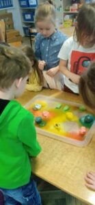 Dzieci stoją przy stoliku ,na którym znajduje się tacka z kolorowymi jajkami uformowanymi z masy. Obserwują rozpuszczanie się sodowych jaj.