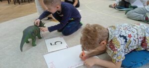 Chłopiec piszący na kartce papieru, w tle drugi chłopiec mierzący dinozaura.