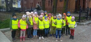 Grupa dzieci w odblaskowych kamizelkach stoi przy pomniku Mikołaja Koprenika.