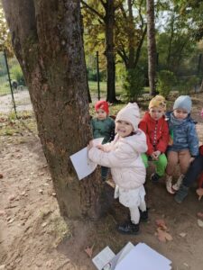 Dziecko przykłada kartkę do pnia drzewa. Z tyłu siedzi na ławce grupa dzieci.