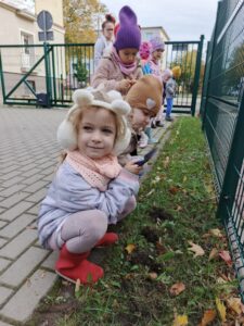 Dziewczynka w nausznikach sadzi kwiaty, w tle grupa dzieci.