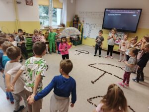 Grupa dzieci stoi w sali przedszkolnej, dziewczynka trzyma parasolkę.