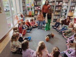 Grupa dzieci siedzi w kole na dywanie, w centrum znajdują się chłopiec, dziewczynka i dorosła kobieta.