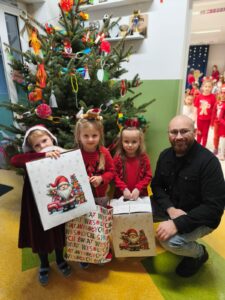 Dzieci z torbami prezentowymi, w tle choinka.