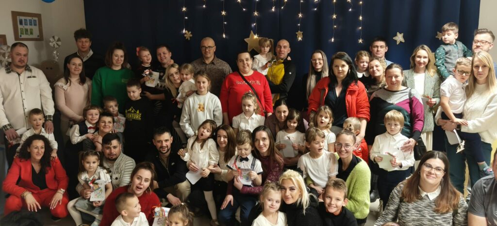 Grupa dzieci i rodziców , w tle granatoe zasłony, dekoracyjne oświetlenie.