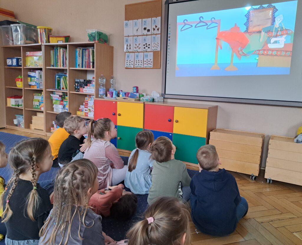 Dzieci siedzą na dywanie i ogladają na tablicy multimedialnej film edukacyjny