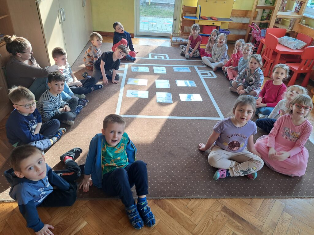 Dzieci siedzą na dywanie, na którym jest przyklejony kontur garnka. W konturze są ułożone ilustracje z symbolami pogody.