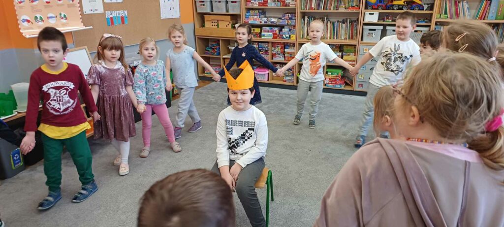 chłopiec siedzi na krześle, dzieci tańczą w kole i śpiewają piosenkę "Sto lat"