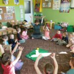 dzieci i nauczycielka siedzą w kole, ilustruja piosenkę, podnoszą ręce do góry