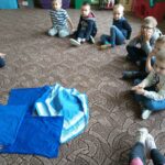dzieci siedza na dywanie. po środki leża niebieskie chusty, pod którymi jest ukruty papier