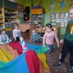 grupa dzieci na dywanie, jedno dziecko jest pod chustą, wyciąga nogę przez dziurę