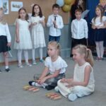 grupa dzieci stoi dokoła sali, przed nimi dwie dziewczynki siedzą i trzymają pałeczki do grania