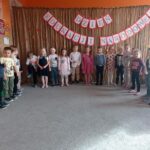Dzieci śpiewają piosenkę ustawione w półkolu na tle dekoracji z okazji Dnia Edukacji Narodowej