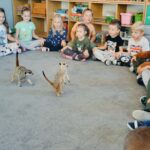Dzieci w kole siedzą na dywanie. W środku koła stoją surykatki