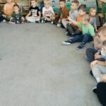 Dzieci w kole siedzą na dywanie. W środku koła stoją surykatki