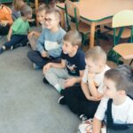 Dzieci siedzą na dywanie, obok biega surykatka