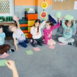 Dzieci siedzą na dywanie i podnoszą różową lub zieloną kartkę.