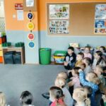 Dzieci siedzą na dywanie. Przed dziećmi stoi policjant