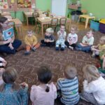 grupa dzieci siedzi z prowadzącą na dywanie i oglądają obrazki