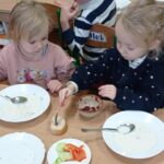 dwie dziewczynki siedzą przy stoliku, jedna smaruje kanapkę