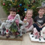 troje dzieci siedzi przed choinką, trzyma w ręku prace wykonane na konkurs