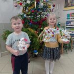 dwoje dzieci siedzi przed choinką, trzyma w ręku prace wykonane na konkurs