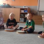 grupa dzieci siedzi na dywanie, przed nimi bębenki