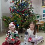 dwie dziewczynki siedzą przed choinką, trzyma w ręku prace wykonane na konkurs