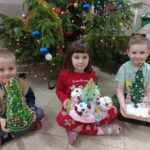 troje dzieci siedzi przed choinką, trzyma w ręku prace wykonane na konkurs