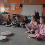 grupa dzieci siedzi na dywanie, przed nimi bębenki